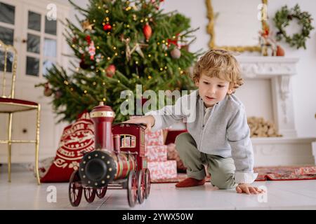 Festliche Weihnachtsszene mit einem kleinen Kind, das mit einem Spielzeugzug in der Nähe eines wunderschön geschmückten Baumes spielt Stockfoto