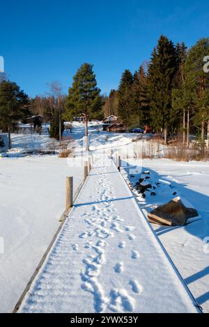 An einem sonnigen Wintertag in Västanvik, Dalarna, zeigt sich ein schneebedecktes Dock, das zu einer unberührten Landschaft führt. Fußspuren zeichnen einen Weg über den Schnee, umgeben Stockfoto