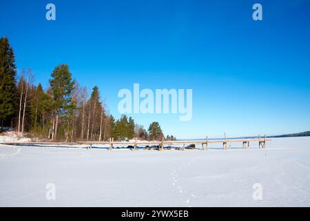 Ein klarer Wintertag in Västanvik, Dalarna, bietet einen ruhigen, schneebedeckten See. Ein rustikaler Holzsteg erstreckt sich über das Eis, umgeben von immergrünem t Stockfoto