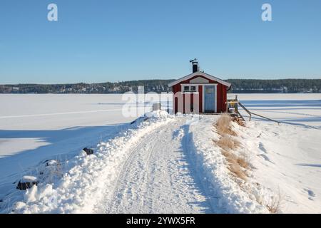 Ein kleines rotes Häuschen steht friedlich auf einem schneebedeckten Pfad, der zu seiner Haustür führt, umgeben von dem ruhigen gefrorenen See und der Winterlandschaft in Västanvik, Stockfoto