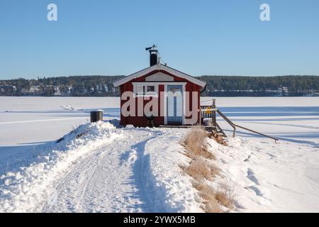 Eine gemütliche rote Hütte liegt allein an einem verschneiten Pfad mit Blick auf einen riesigen gefrorenen See in Västanvik, Dalarna. Die ruhige Winterlandschaft wird durch cl hervorgehoben Stockfoto