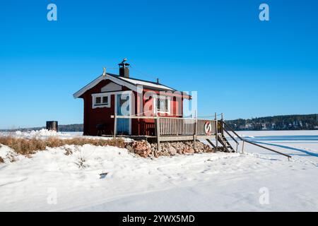 Eine bezaubernde rote Hütte steht in der Nähe eines gefrorenen Sees in Västanvik, Dalarna. Schnee bedeckt den Boden und der klare blaue Himmel rundet die ruhige Winterlandschaft ab Stockfoto