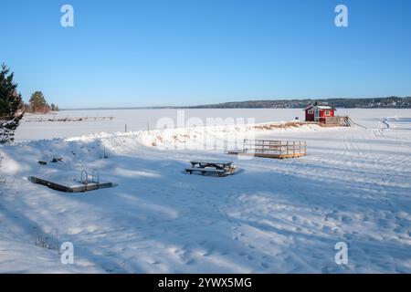 Ein beschaulicher Wintertag in Västanvik, Dalarna, mit einer Schneedecke, die die Landschaft bedeckt. Der gefrorene See reflektiert den klaren blauen Himmel, während ein rotes c Stockfoto