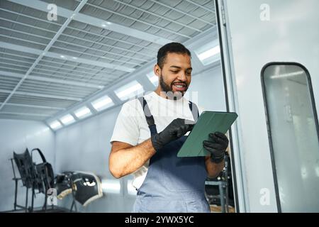 Ein gutaussehender junger Mechaniker liest bei Tageslicht in der geschäftigen Werkstatt auf einem Tablet die Jobdetails. Stockfoto