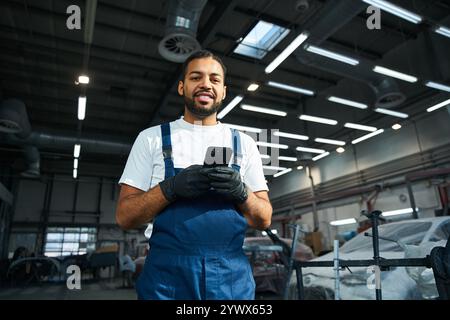 Junger Mechaniker in blauen Overalls benutzt Smartphone, während er in der Werkstatt von Fahrzeugen umgeben ist. Stockfoto