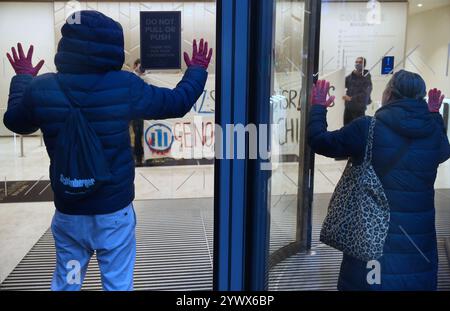 Bimingham, England, Großbritannien. Dezember 2024. Aktivisten protestieren im Empfangsbereich des Bürogebäudes, während Unterstützer während der Demonstration ihre rot bedeckten Hände auf das Glas drücken. Pro-palästinensische Aktivisten zielen auf die Versicherungsbüros der Allianz in ihrem Colmore Building in Birmingham, die den Empfangsbereich besetzen und draußen protestieren. Sie zielen auf die Allianz ab, weil sie das israelische Rüstungsunternehmen Elbit Systems versichern und in das israelische Rüstungsunternehmen Elbit Systems investieren und entschlossen sind, den Druck des direkten Handelns aufrecht zu erhalten, damit es zu einem großen Nachteil wird, mit ihnen zusammenzuarbeiten. Sie verlangen, dass die Firma Elbit Systems abwirft. Stockfoto