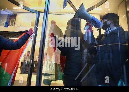 Bimingham, England, Großbritannien. Dezember 2024. Aktivisten protestieren im Empfangsbereich des Bürogebäudes, während Unterstützer, die in der Drehtür eingeschlossen sind, während der Demonstration das Chanten aufrechterhalten. Pro-palästinensische Aktivisten zielen auf die Versicherungsbüros der Allianz in ihrem Colmore Building in Birmingham, die den Empfangsbereich besetzen und draußen protestieren. Sie zielen auf die Allianz ab, weil sie das israelische Rüstungsunternehmen Elbit Systems versichern und in das israelische Rüstungsunternehmen Elbit Systems investieren und entschlossen sind, den Druck des direkten Handelns aufrecht zu erhalten, damit es zu einem großen Nachteil wird, mit ihnen zusammenzuarbeiten. Sie verlangen, dass die Firma Elbit abwirft Stockfoto