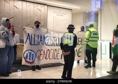 Bimingham, England, Großbritannien. Dezember 2024. Aktivisten protestieren im Empfangsbereich des Bürogebäudes mit einem Banner mit der Aufschrift „˜Allianz sichert Israels Völkermord-Maschine“ während der Demonstration, während Polizeibeamte anwesend sind und sie zum Verlassen bringen. Pro-palästinensische Aktivisten zielen auf die Versicherungsbüros der Allianz in ihrem Colmore Building in Birmingham, die den Empfangsbereich besetzen und draußen protestieren. Sie zielen auf die Allianz ab, weil sie das israelische Rüstungsunternehmen Elbit Systems versichern und in das israelische Rüstungsunternehmen Elbit Systems investieren und entschlossen sind, den Druck des direkten Handelns aufrecht zu erhalten, damit es zu einem großen Nachteil für wird Stockfoto