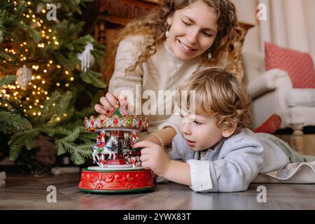 Mutter und Sohn richten während der Weihnachtszeit gemeinsam ein Weihnachtskarussell in einem gemütlichen Wohnzimmer ein Stockfoto