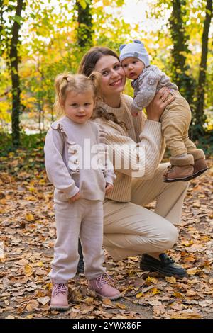 Lächelnde Mutter auf einem Spaziergang in einem Herbstpark mit ihrem neugeborenen Jungen und ihrer kleinen Tochter. Glückliche Familie. Mutterschaft. Stockfoto