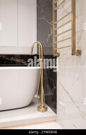 Langer stilvoller Goldhahn auf dem Boden in der Nähe der weißen Badewanne auf Granitwand Hintergrund. Moderne Badezimmer-Innenausstattung. Stockfoto