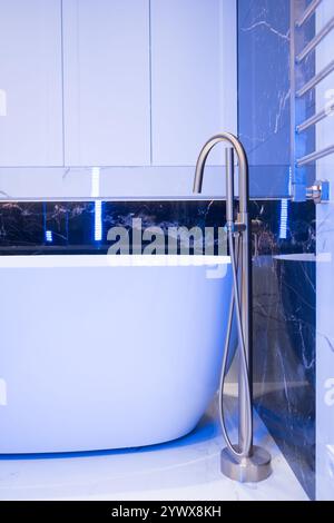 Langer stilvoller Goldhahn auf dem Boden in der Nähe der weißen Badewanne auf Granitwand Hintergrund. Moderne Badezimmer-Innenausstattung. Stockfoto