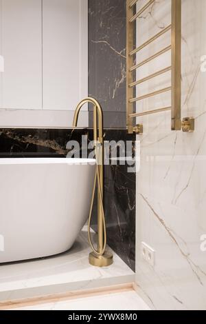 Langer stilvoller Goldhahn auf dem Boden in der Nähe der weißen Badewanne auf Granitwand Hintergrund. Moderne Badezimmer-Innenausstattung. Stockfoto