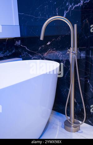 Langer stilvoller Goldhahn auf dem Boden in der Nähe der weißen Badewanne auf Granitwand Hintergrund. Moderne Badezimmer-Innenausstattung. Stockfoto