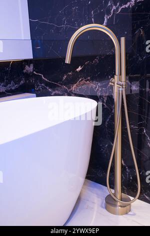 Langer stilvoller Goldhahn auf dem Boden in der Nähe der weißen Badewanne auf Granitwand Hintergrund. Moderne Badezimmer-Innenausstattung. Stockfoto