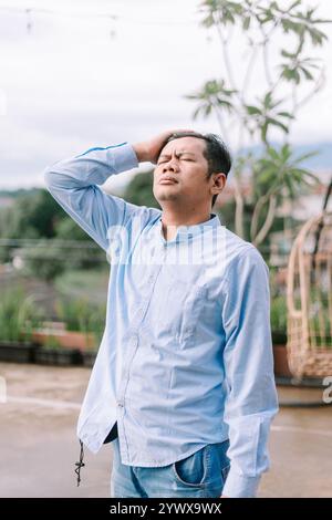 Ein südostasiatischer Mann, der seinen Kopf unbehaglich hält, sichtbar Kopfschmerzen erlebt, einen Moment körperlicher Belastung oder Stress festnimmt. Stockfoto