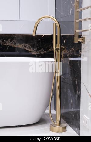 Langer stilvoller Goldhahn auf dem Boden in der Nähe der weißen Badewanne auf Granitwand Hintergrund. Moderne Badezimmer-Innenausstattung. Stockfoto