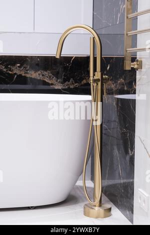 Langer stilvoller Goldhahn auf dem Boden in der Nähe der weißen Badewanne auf Granitwand Hintergrund. Moderne Badezimmer-Innenausstattung. Stockfoto