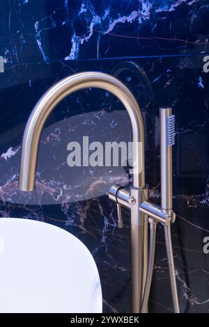 Langer stilvoller Goldhahn auf dem Boden in der Nähe der weißen Badewanne auf Granitwand Hintergrund. Moderne Badezimmer-Innenausstattung. Stockfoto