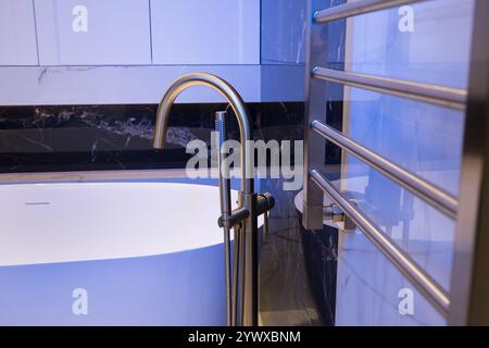 Langer stilvoller Goldhahn auf dem Boden in der Nähe der weißen Badewanne auf Granitwand Hintergrund. Moderne Badezimmer-Innenausstattung. Stockfoto