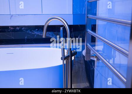 Langer stilvoller Goldhahn auf dem Boden in der Nähe der weißen Badewanne auf Granitwand Hintergrund. Moderne Badezimmer-Innenausstattung. Stockfoto