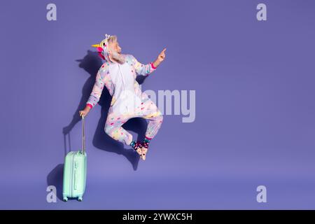Reifer Mann im Einhorn-Pyjama, der fröhlich mit Koffer vor leuchtendem lila Hintergrund springt Stockfoto