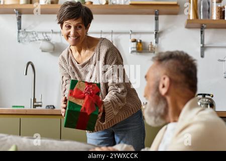 Reife Frau präsentiert ihrem lächelnden Partner in einem gemütlichen Wohnbereich ein wunderschön verpacktes Geschenk. Stockfoto