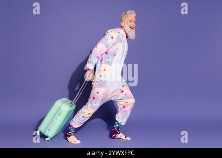 Fröhlicher Mann im bunten Einhorn-Pyjama mit einem Koffer vor violettem Hintergrund Stockfoto