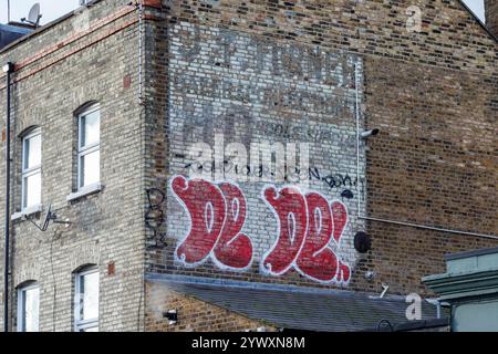 Ein Geisterschild mit der Werbung „J. T. Turner, Baker & Confectioner, Hovis, Schools Supported“ auf einer Ziegelwand mit Giebelenden, bedeckt mit Graffiti, London, Großbritannien Stockfoto