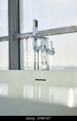 Nahaufnahme: Verchromter Wasserhahn im Badezimmer mit fließendem Wasser über moderner freistehender Badewanne im minimalistischen Badezimmer, Kopierraum Stockfoto