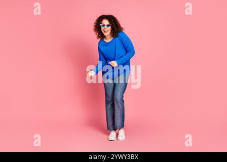 Fröhliche junge Frau mit lockigem Haar in einem lässigen blauen Pullover tanzt vor einem rosa Hintergrund und fördert Mode und Spaß. Stockfoto