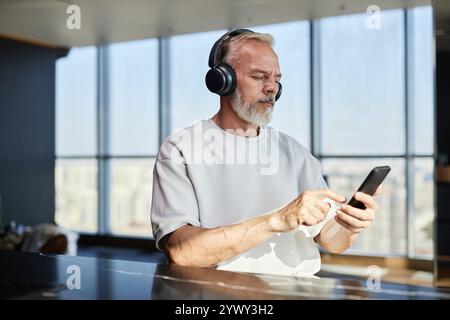 Mittelgroße Aufnahme eines ernsten, Reifen Mannes mit ordentlichem Bart, der das Smartphone benutzt und Musik über Kopfhörer hört, während er am Tisch in einer modernen Wohnung sitzt, Kopierraum Stockfoto