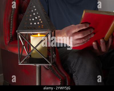 Nahaufnahme eines Mannes, der in einem traditionellen Ledersessel sitzt, ein altes gebundenes Buch hält und bei Kerzenschein liest. Stockfoto