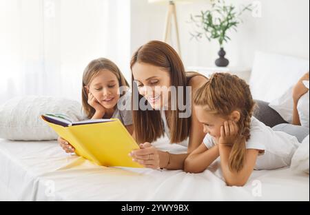 Lächelnde Mutter und ihre beiden Kinder liegen auf dem Bett, lesen zusammen Buch und genießen das Erzählen von Geschichten. Stockfoto