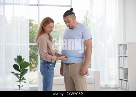 Ernährungswissenschaftler, der die Taille eines übergewichtigen Mannes mit Klebeband in der Klinik misst Stockfoto
