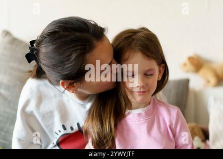 Mutter küsst ihre kleine Tochter auf dem Sofa in der Wohnung. Konzept gesunder Familienbeziehungen Stockfoto
