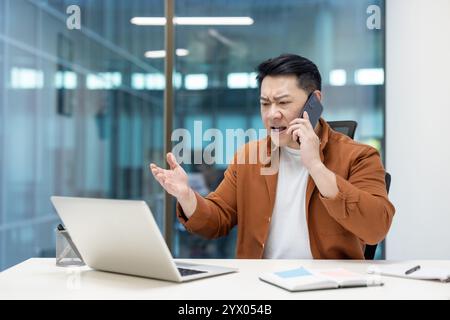 Asiatischer Mann reifer Geschäftsmann, der während eines Telefonanrufs Frustration ausdrückt, während er in seinem Büro arbeitet. Stockfoto