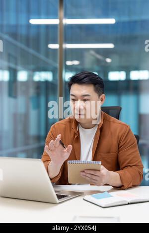 Asiatischer Mann reifer Geschäftsmann, der an einem modernen Arbeitsplatz arbeitet, Notizen macht und online kommuniziert. Repräsentiert Produktivität und digitale Arbeitsumgebungen. Stockfoto