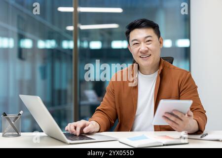 Asiatischer Mann reifer Geschäftsmann, der in einem Büro mit Tablet und Laptop arbeitet und selbstbewusst lächelt. Die Atmosphäre spiegelt Produktivität, Erfolg und eine positive Einstellung wider. Stockfoto