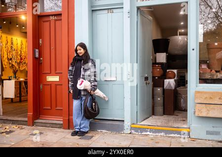 London, Großbritannien, 1. Dezember 2024, posiert Eine junge Asiatin in einer karierten Wolljacke mit einem Blumenstrauß neben einer bunten Wand auf dem Columbia Flower Market. Stockfoto