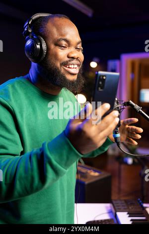 Fröhlicher Musiker, der seine neue musikalische Kreation am Mikrofon singt und Audiodateien aufnimmt, um sie in der Postproduktion zu bearbeiten. Junger Künstler Songwriter liest die kreativen Verse und Elemente am Telefon. Stockfoto