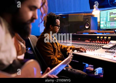 Talentierter Sänger Komponist, der Gitarrennoten in professionellen Musikstudios aufnimmt und elektroakustische Klänge verwendet. Künstler, der mit einem Tontechniker im Kontrollraum arbeitet, Postproduktion. Stockfoto