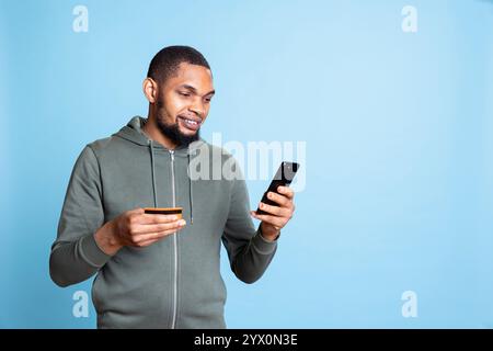 afroamerikanischer Shopaholic, der Kreditkartennummern eingibt, um eine Zahlung auf seinem Smartphone zu tätigen und Dinge zu kaufen. Fröhlicher, aufgeregter Kerl macht Online-Shopping, E-Commerce. Stockfoto