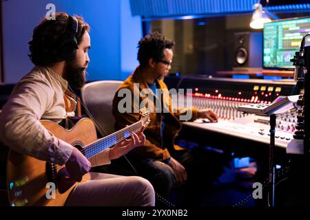 Sänger spielt einen neuen Hit auf seiner Gitarre im Kontrollraum, nimmt Tracks auf und komponiert sie mit Sounddesigner im professionellen Musikstudio. Künstler, der auf einem elektroakustischen Instrument auftritt. Stockfoto