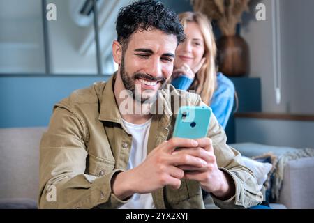 Mann, der ein Smartphone benutzt, während er zu Hause mit einer Frau im Hintergrund sitzt. Stockfoto