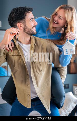 Glückliches Paar, das in gemütlicher Umgebung ein neues Zuhause mit Schlüsseln und Hausmodell feiert. Stockfoto