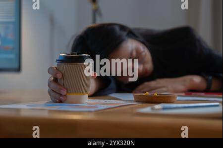 Überarbeitete und müde Geschäftsfrau, die schläft, während sie im Büro auf einem Schreibtisch eine Tasse Kaffee überreicht. Überstundenkonzept. Selektiver Fokus. Stockfoto
