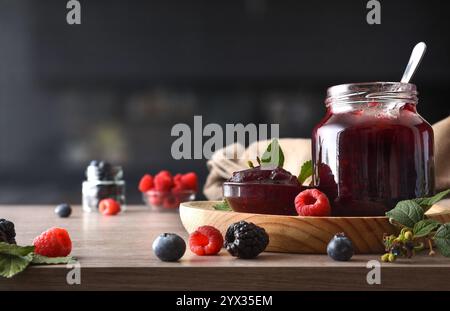 Waldfruchtmarmelade in Glasgläsern auf einer Holzbank mit Beeren in einer schwarzen Küche. Vorderansicht. Stockfoto