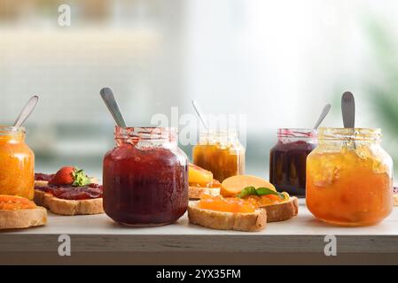 Gläser voller Marmelade mit verschiedenen Geschmacksrichtungen und Brotscheiben mit Marmelade dekoriert und Küchenhintergrund. Vorderansicht. Stockfoto