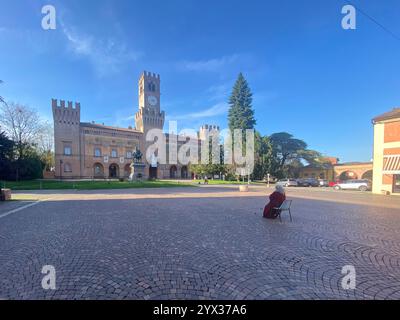 Busseto, Parma, Italien - 23. November 2024 eine ältere Frau sitzt auf der mit Kopfsteinpflaster gepflasterten piazza giuseppe verdi und betrachtet die historische rocca pallavicina Stockfoto